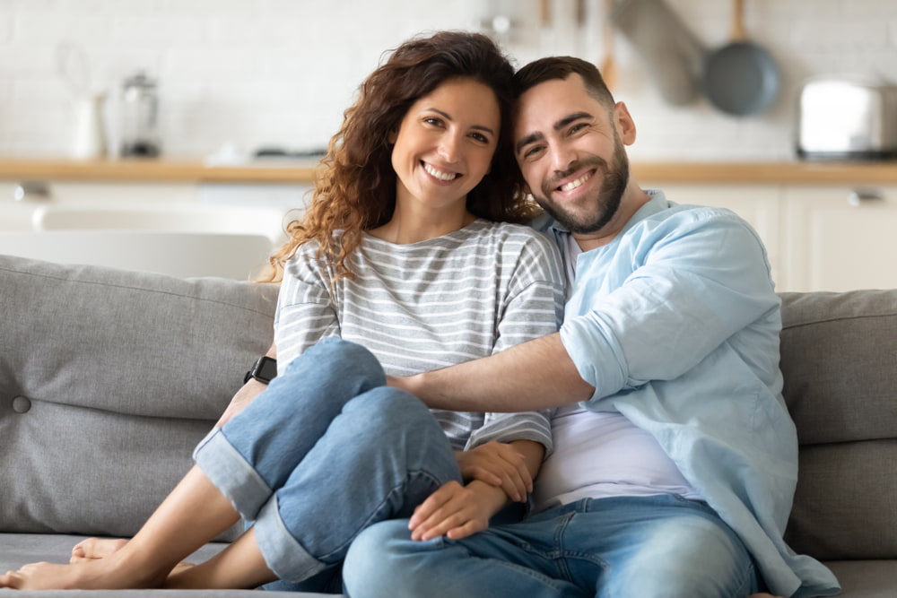 Casal hétero e branco sentados em um sofá e sorrindo.